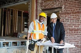 Two men in hard hats and safety vests standing next to a table.