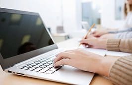 A person typing on a laptop with a pen and paper.