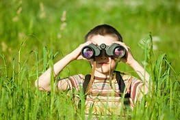 A boy is sitting in the grass looking through binoculars.